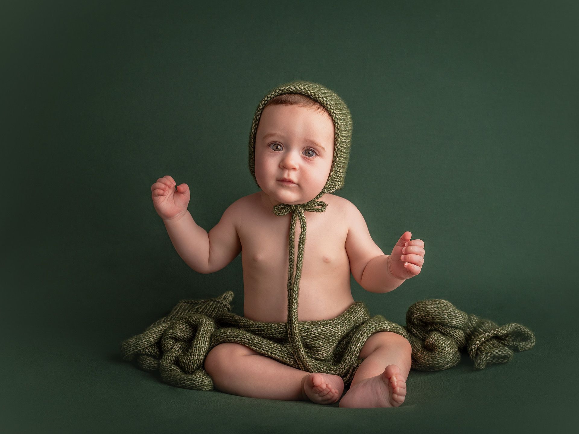 Bebé con gorro verde sentado en estudio, fotografía infantil con fondo liso y cálido.